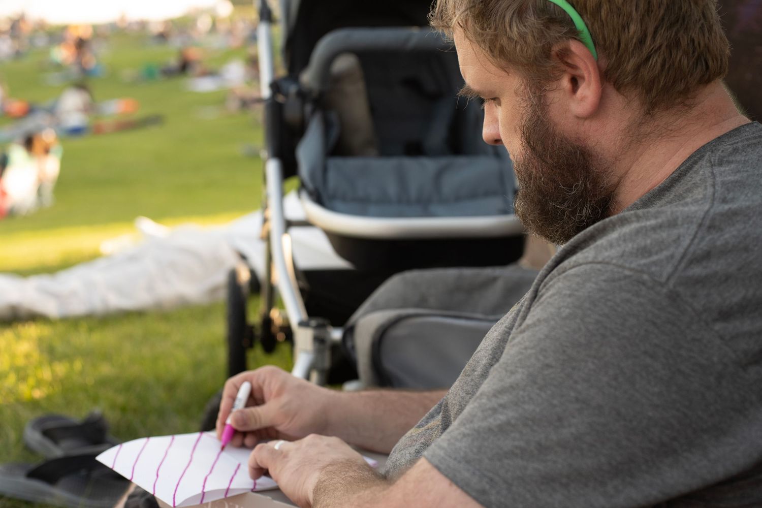 Someone in a gray shirt writes in a notebook outdoors with a stroller visible in the background on a sunny day.