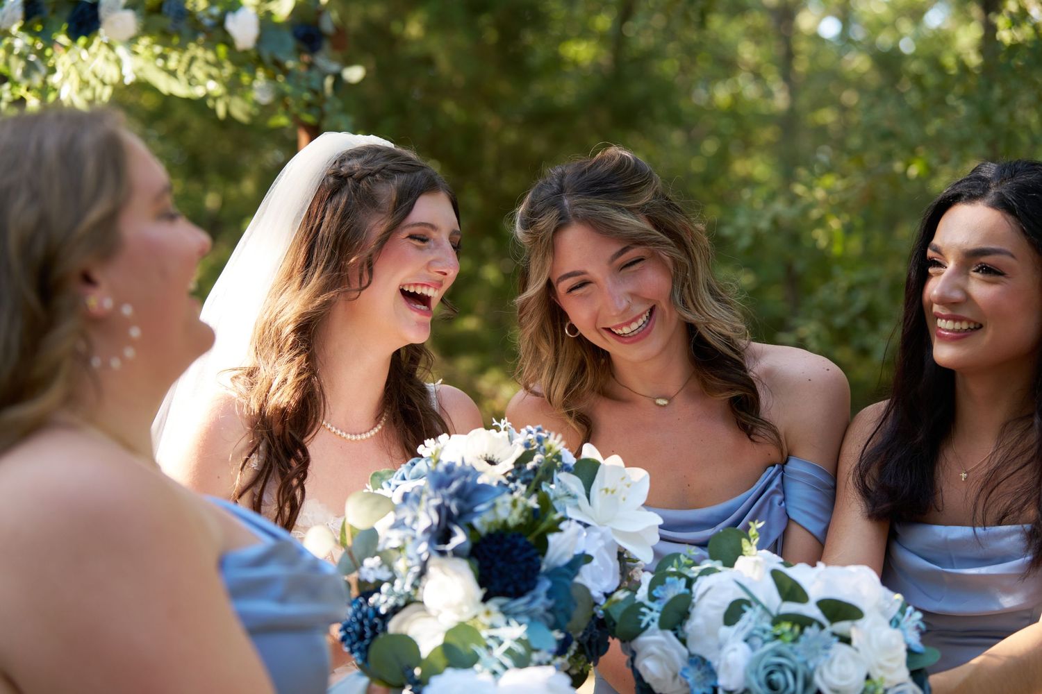 Bridesmaids in light blue dresses laugh together while holding blue and white floral bouquets outdoors.