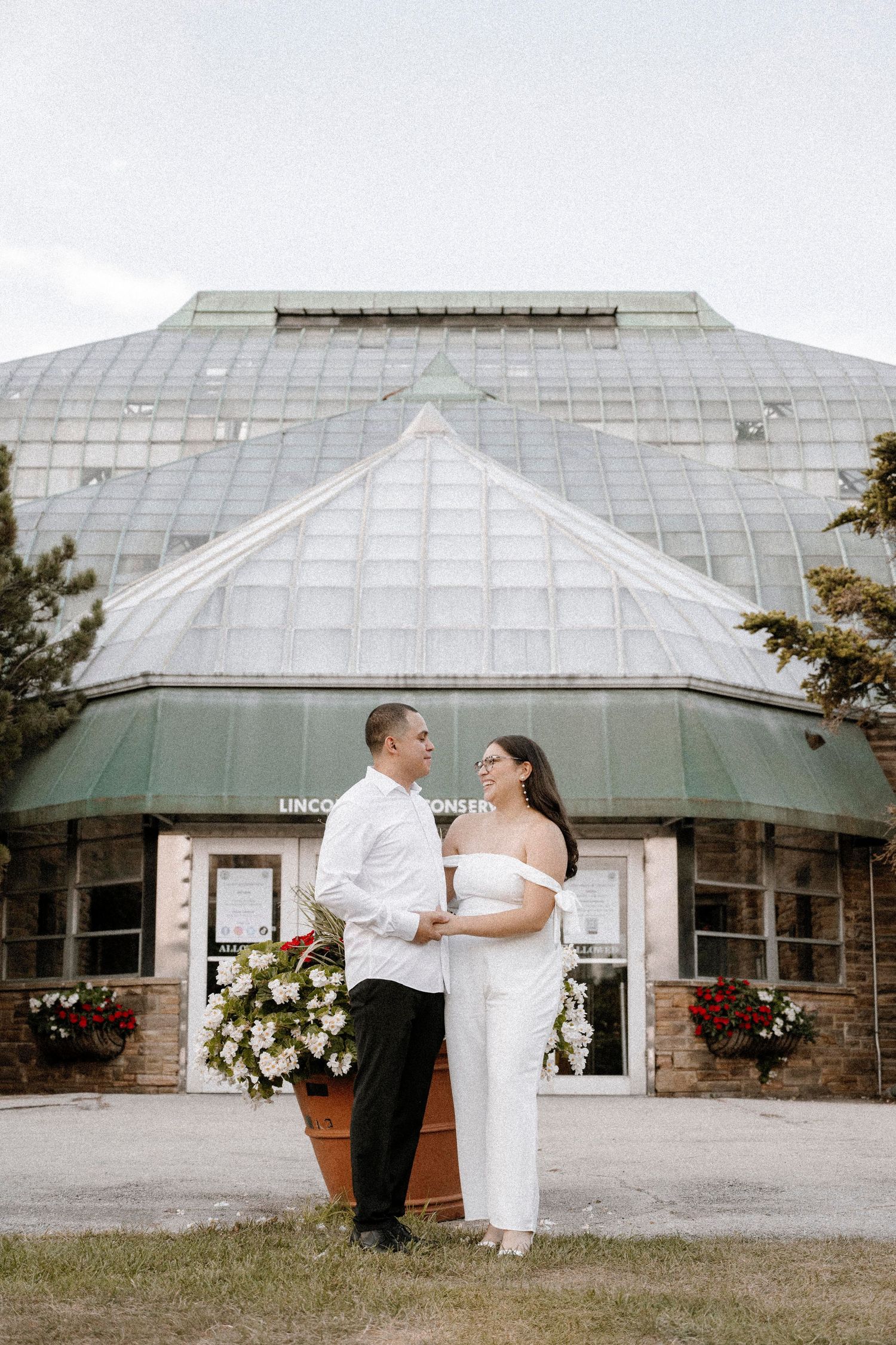 A couple poses together in front of a large glass greenhouse dome decorated with red potted flowers.