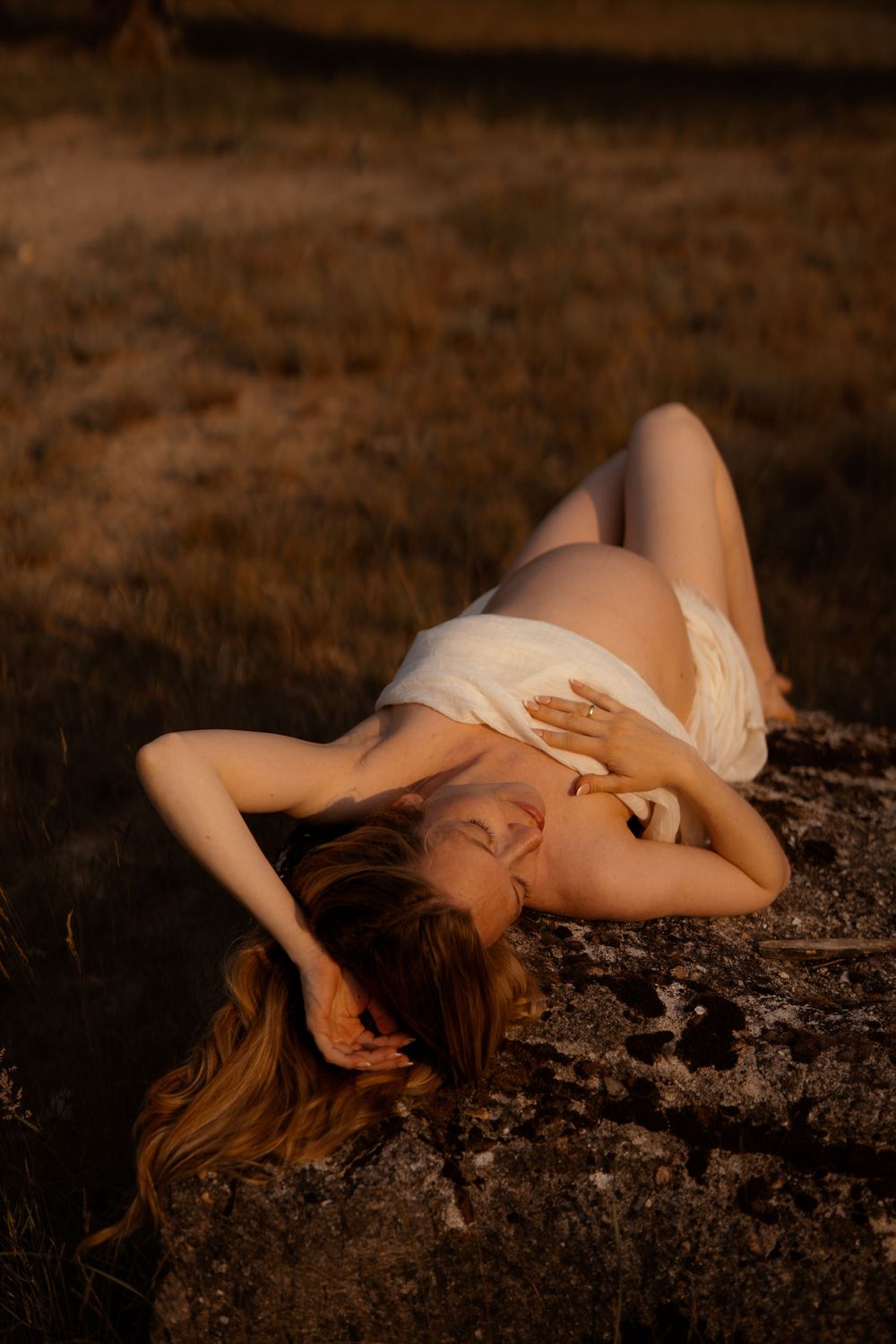 A dreamy portrait at golden hour features a reclining figure on a stone surface wearing ethereal white fabric.