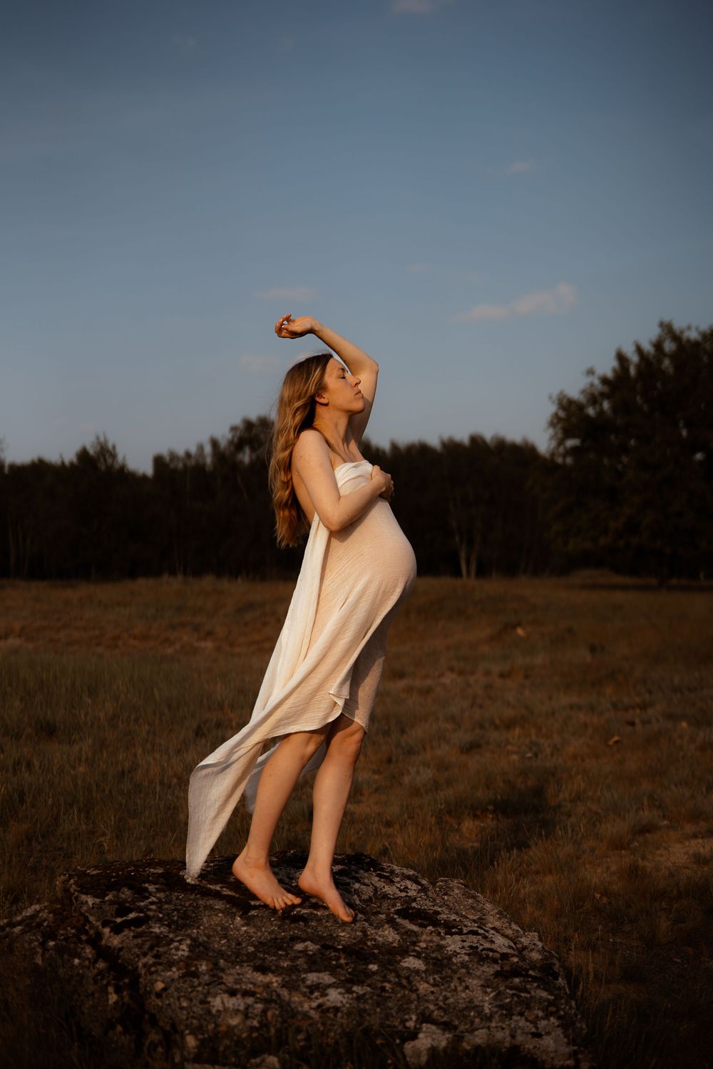 Pregnant woman in flowing cream dress poses gracefully on rock at golden hour in nature setting.