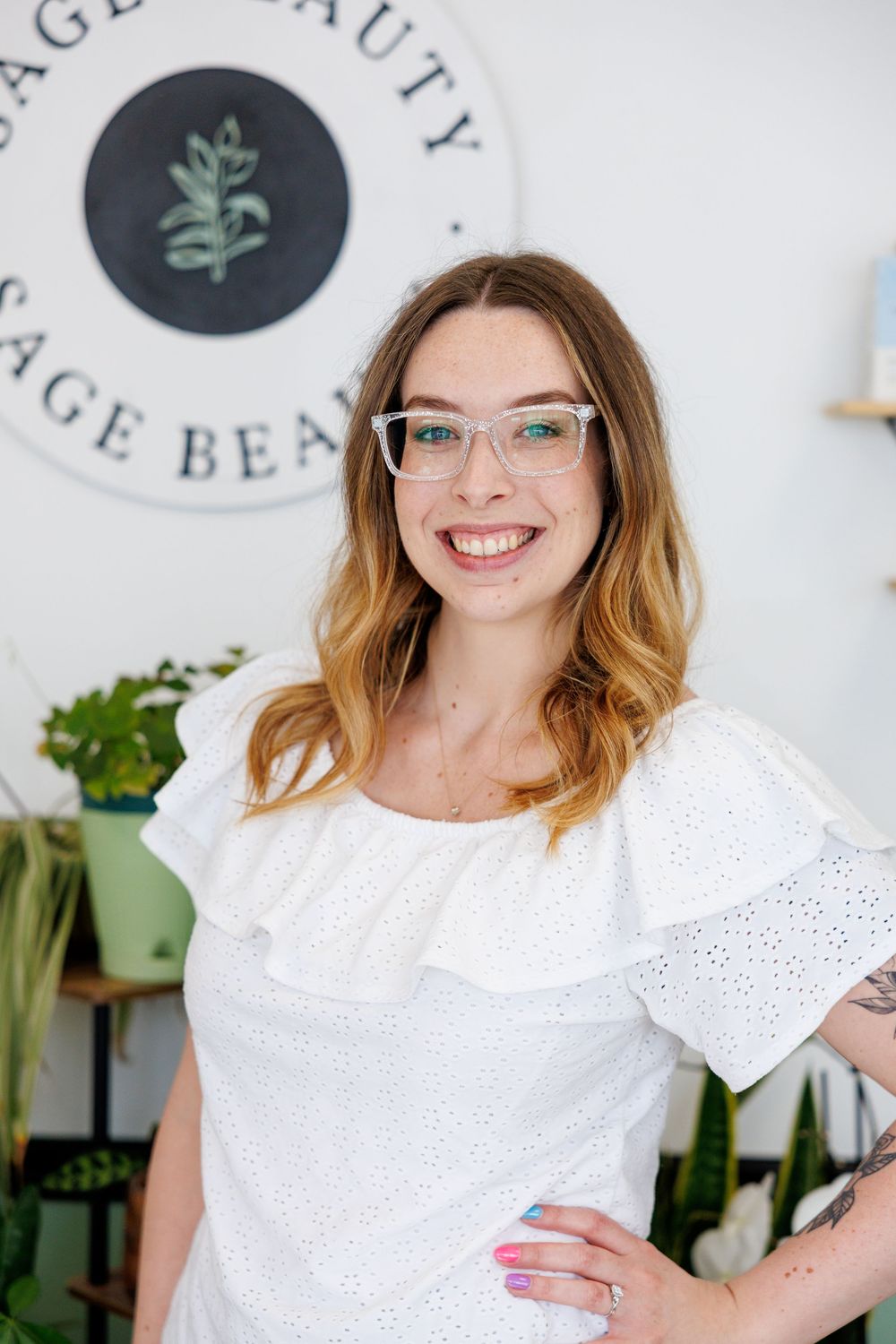 Professional portrait of someone wearing glasses and a white eyelet lace top, standing in a beauty spa or salon space.