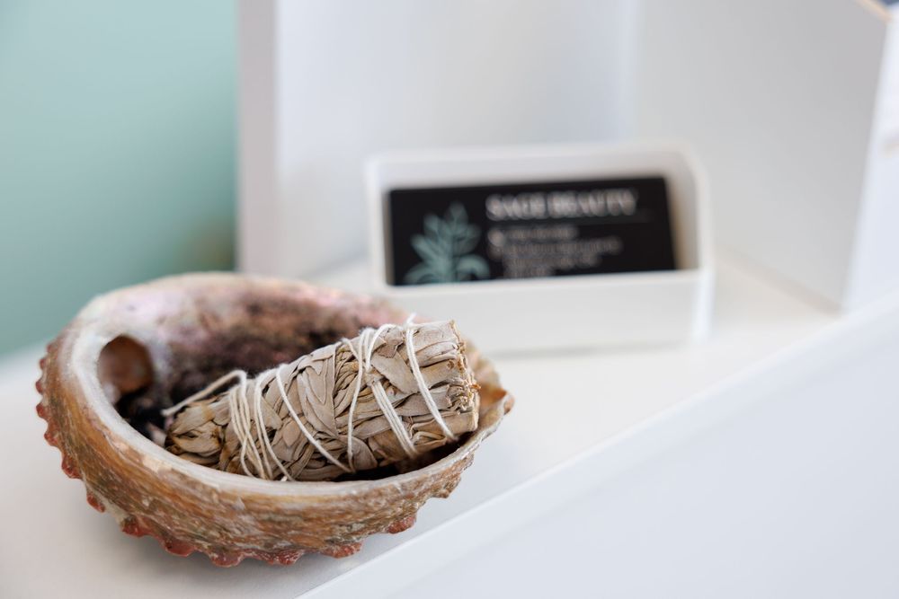 A white sage smudge stick rests in an abalone shell with a box in the blurred background.