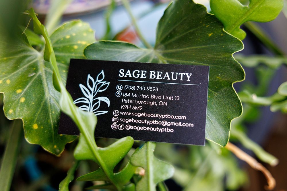 Black business card for Sage Beauty company displayed against green leafy plant background.