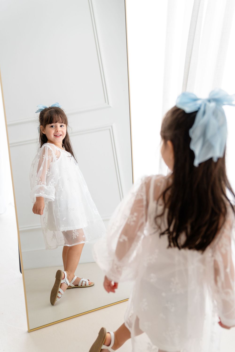 A white lace dress reflection in a full-length mirror with a blue bow hair accessory.