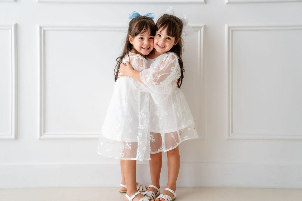 Two young children in matching white lace dresses hug and smile while standing against a white wall.