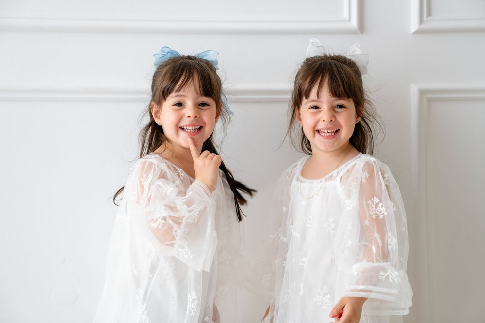 Two young children in white lace dresses laugh and dance together in a bright interior setting.