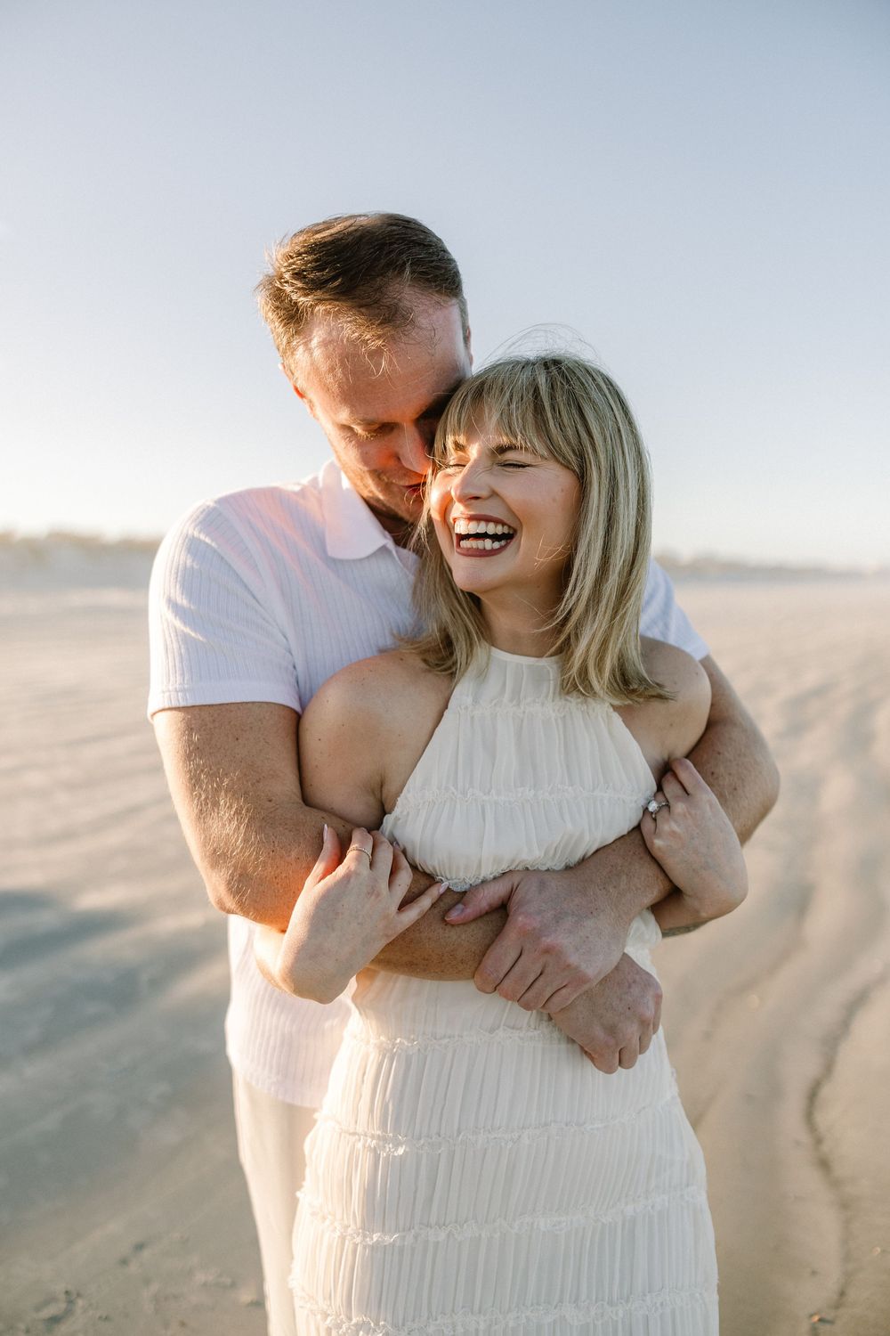 A couple shares an intimate laughing moment together on the beach in evening light.