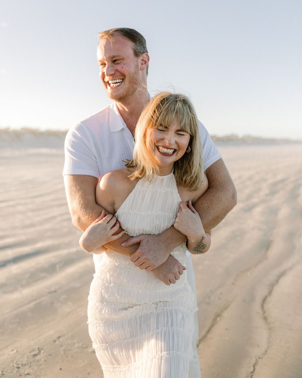 A couple shares a candid laughing moment together on a sandy beach during sunset.