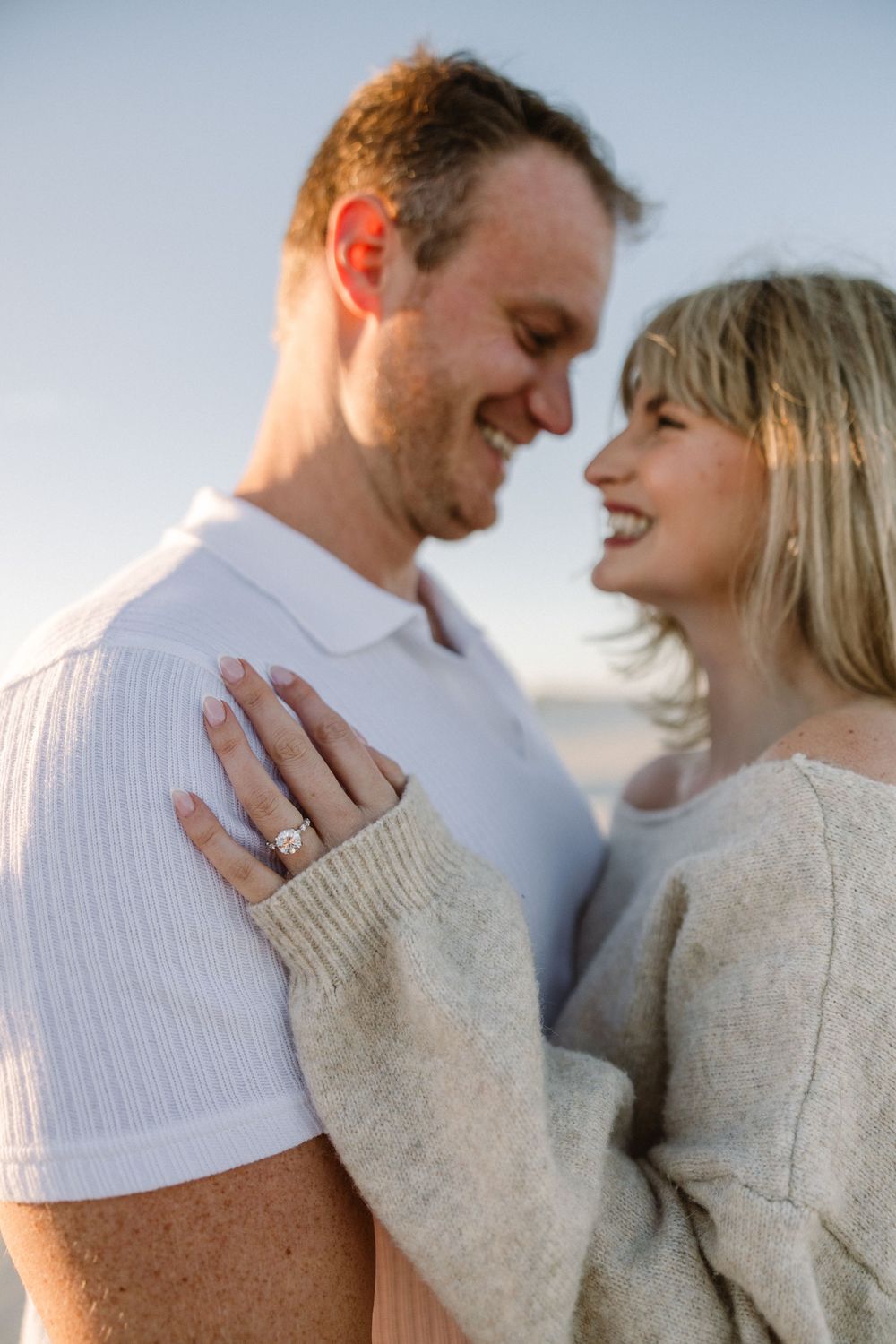 A couple in casual attire shares a romantic moment on the beach while showcasing an engagement ring.