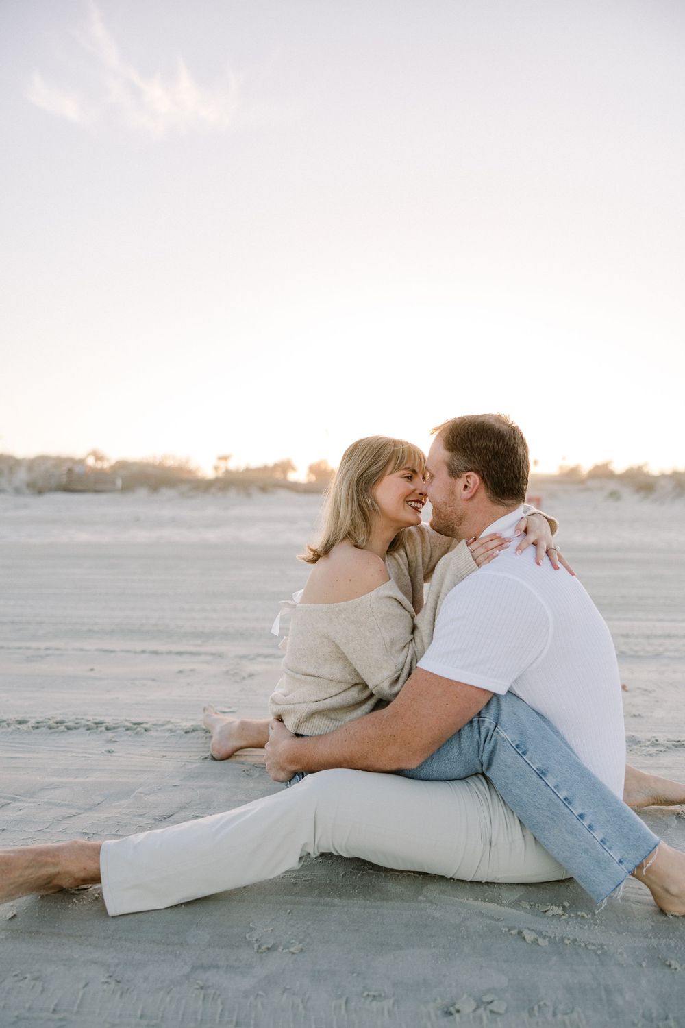 A couple sits together intimately on the beach during sunset in casual attire.