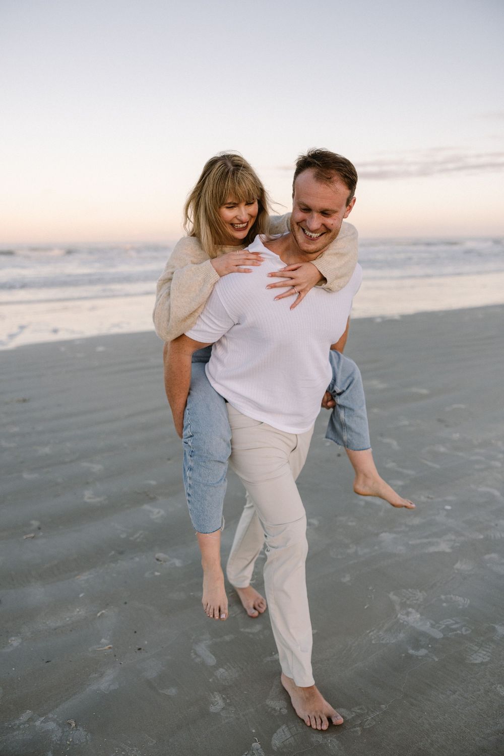 A couple playfully embraces on a beach at sunset while wearing casual white and denim outfits.
