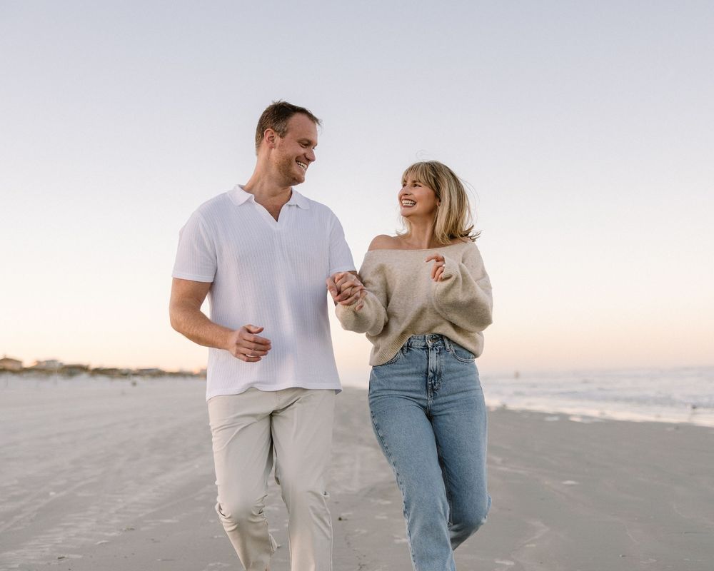 A couple walking and laughing together on the beach at sunset in casual attire.