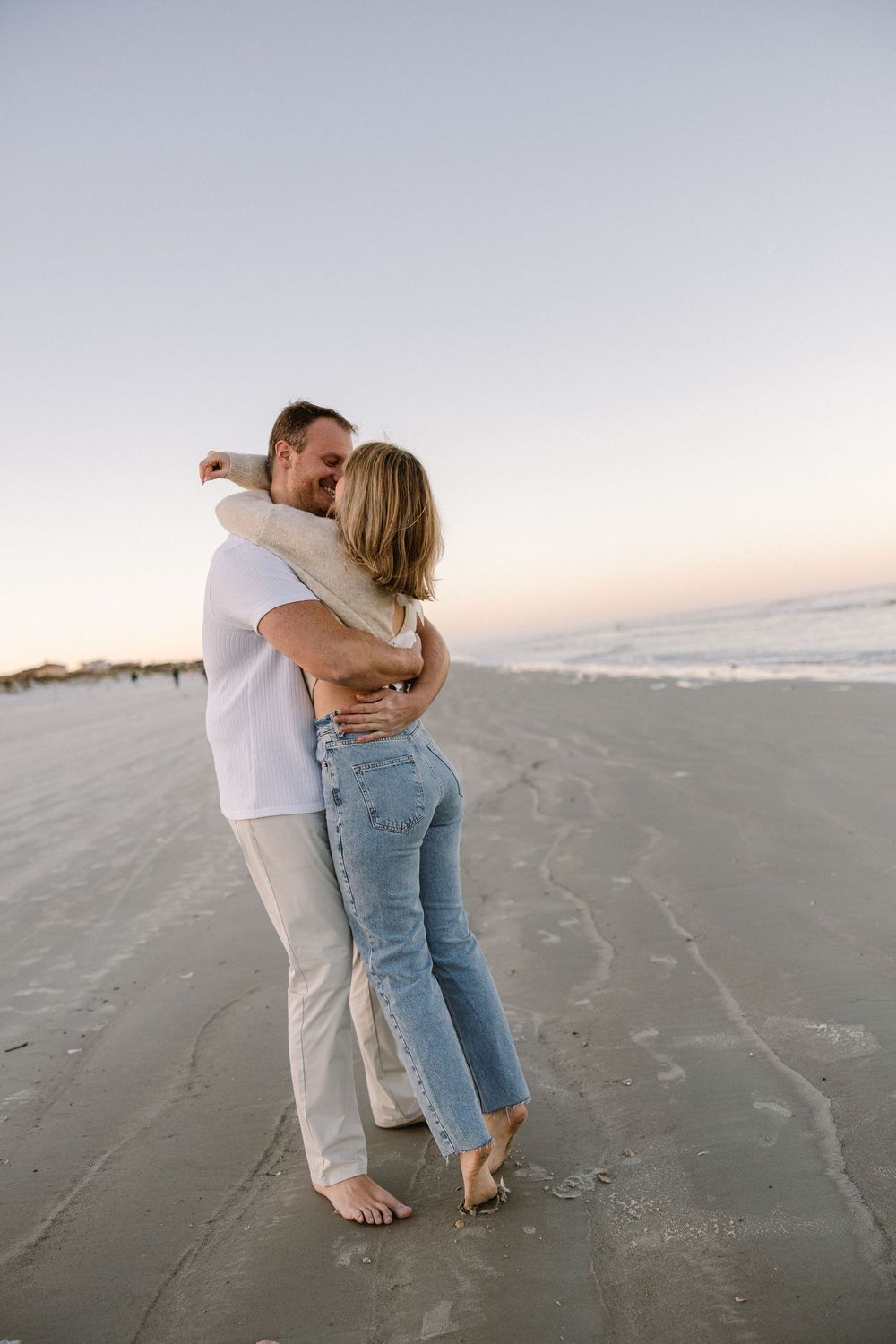 A romantic couple shares an intimate embrace while standing barefoot on a beach at dusk.