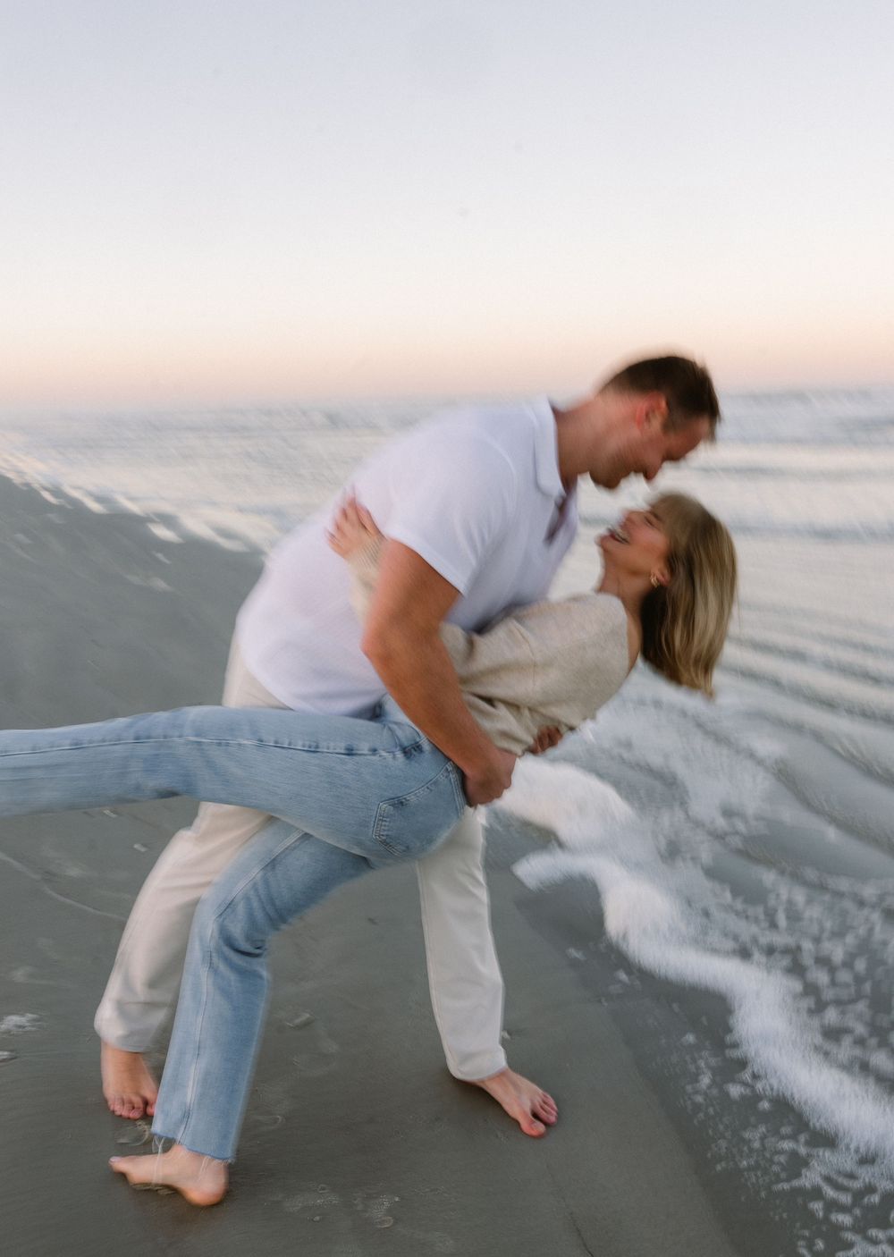 A couple shares a playful dip pose together on the beach with waves in the background.
