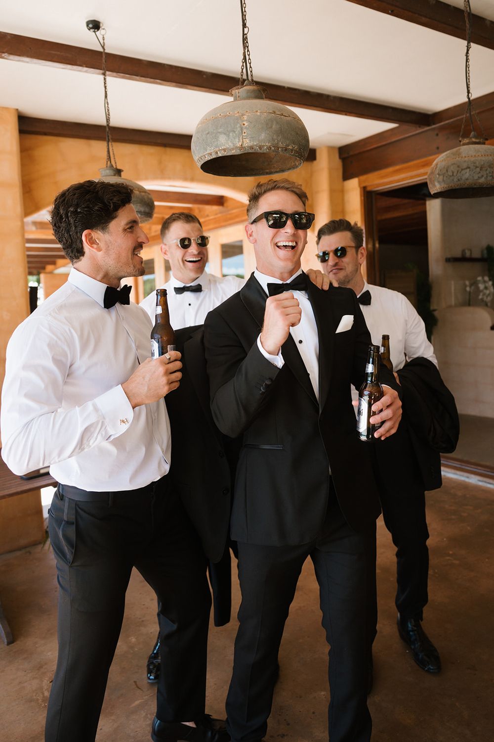 Groomsmen in formal wear sharing drinks and laughing together at a wedding venue with industrial pendant lights.