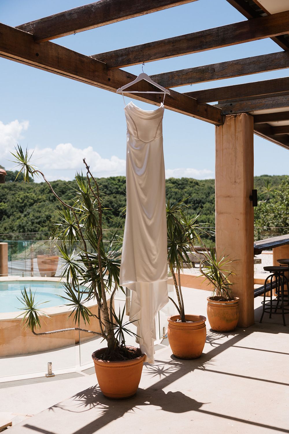 An elegant white wedding dress hangs from wooden beams on a patio with potted palms and a swimming pool view in the background.