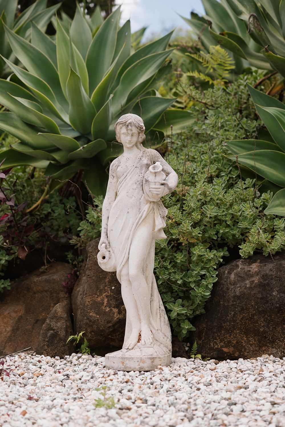 A classical stone garden statue surrounded by large agave plants and succulent landscaping on white gravel ground.
