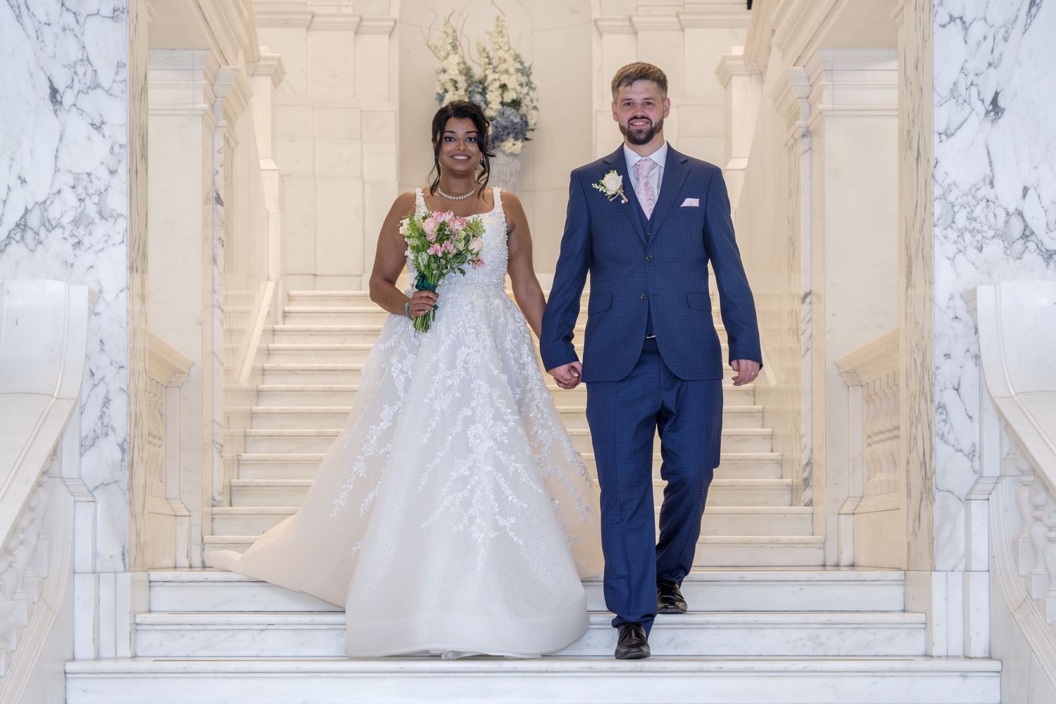 A bride and groom descend a grand marble staircase while holding hands at an elegant wedding venue.