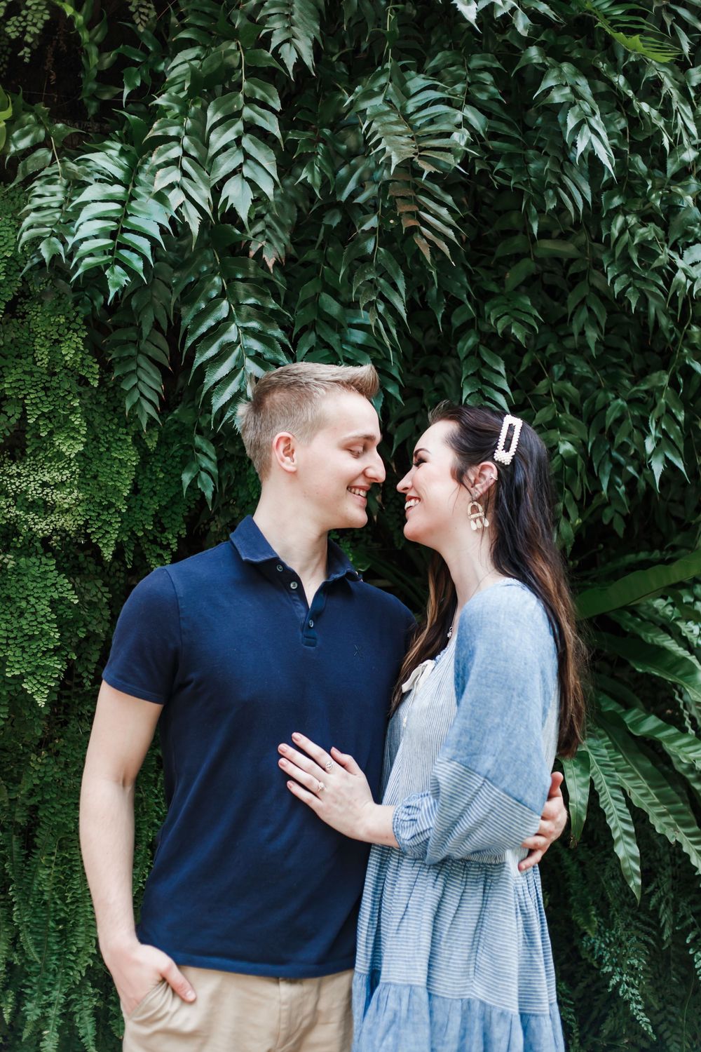 A couple shares a tender moment in front of lush green monstera leaves wearing navy and light blue outfits.