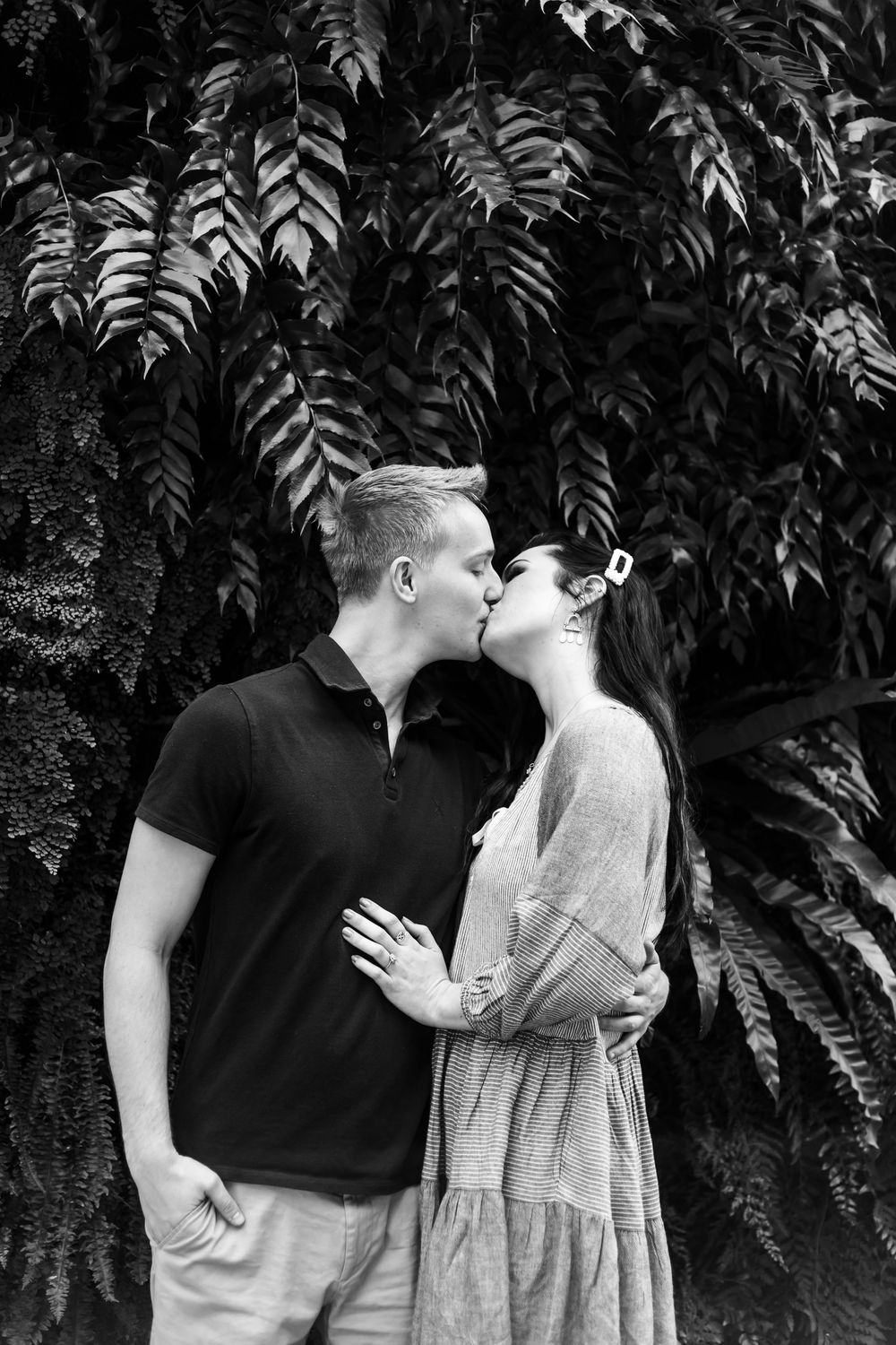 Romantic couple sharing a tender kiss against a backdrop of lush ferns in black and white photography.