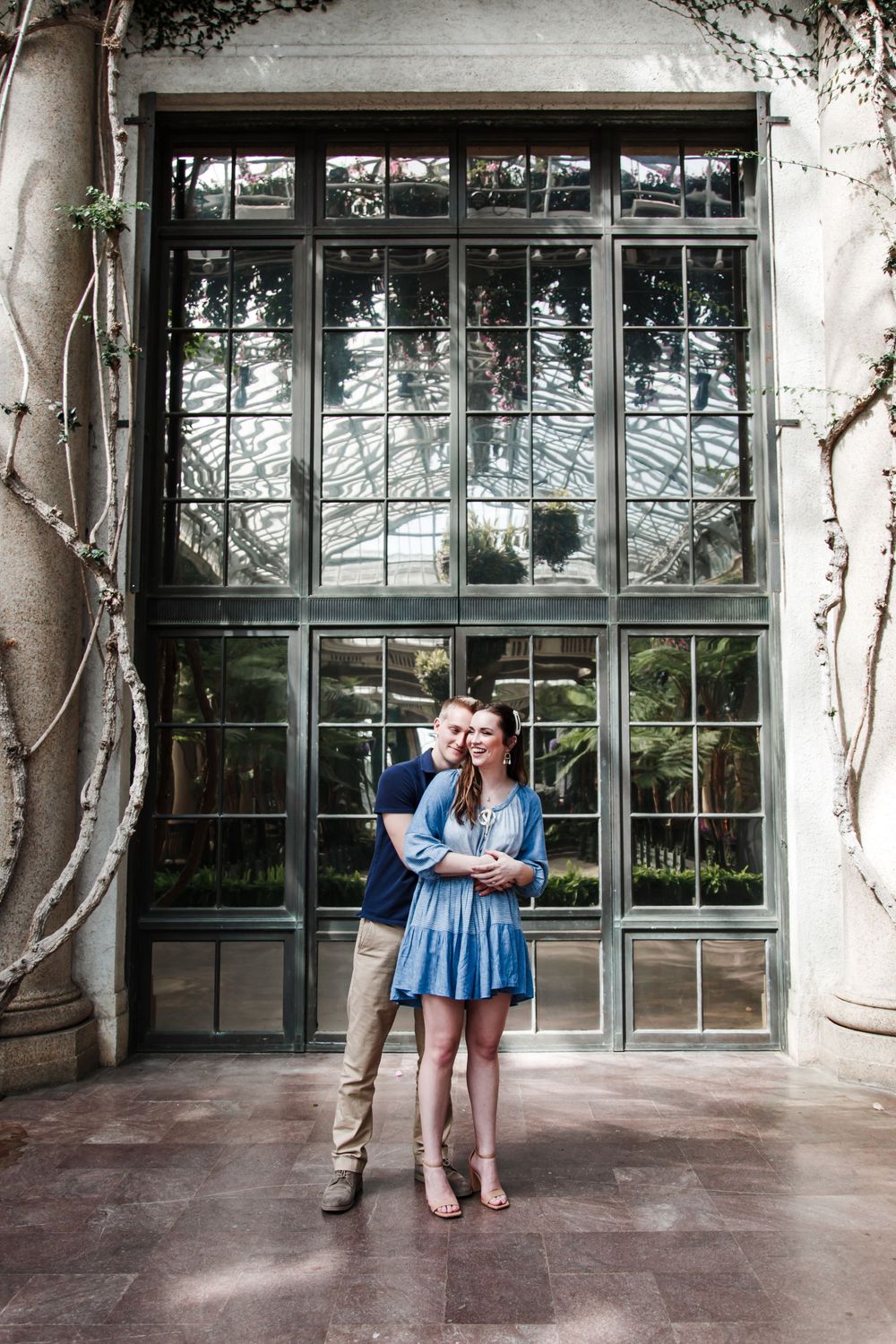 A couple embraces in front of a large industrial window with vines growing along the walls.