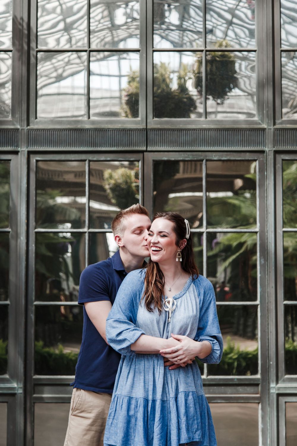A romantic couple embraces in front of a glass greenhouse wall on a sunny day.
