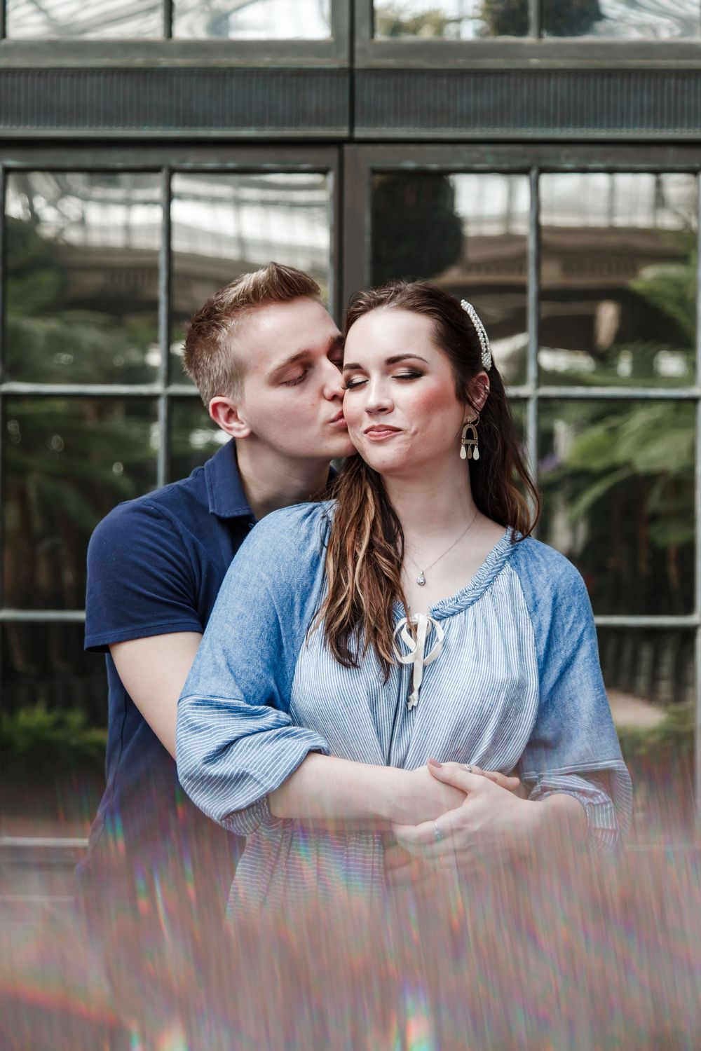 Intimate moment captured between couple near greenhouse windows with soft natural lighting.