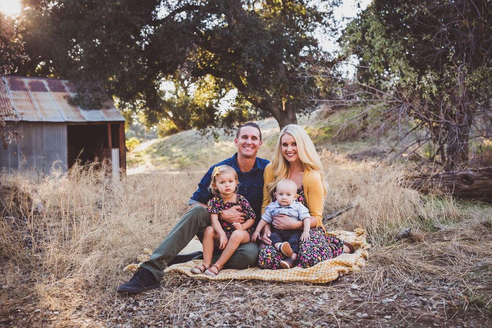 Family sitting together on blanket outdoors in rural setting with tall grass and trees at golden hour.