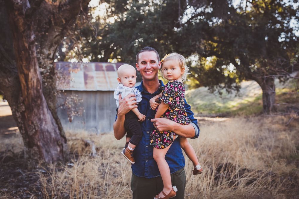 A parent and two young children pose together outdoors in a natural setting with trees during sunset.