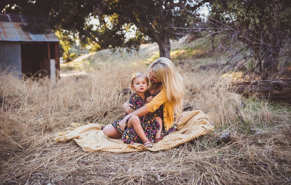 Mother and child sitting on a blanket in golden field near rustic shed during sunset.