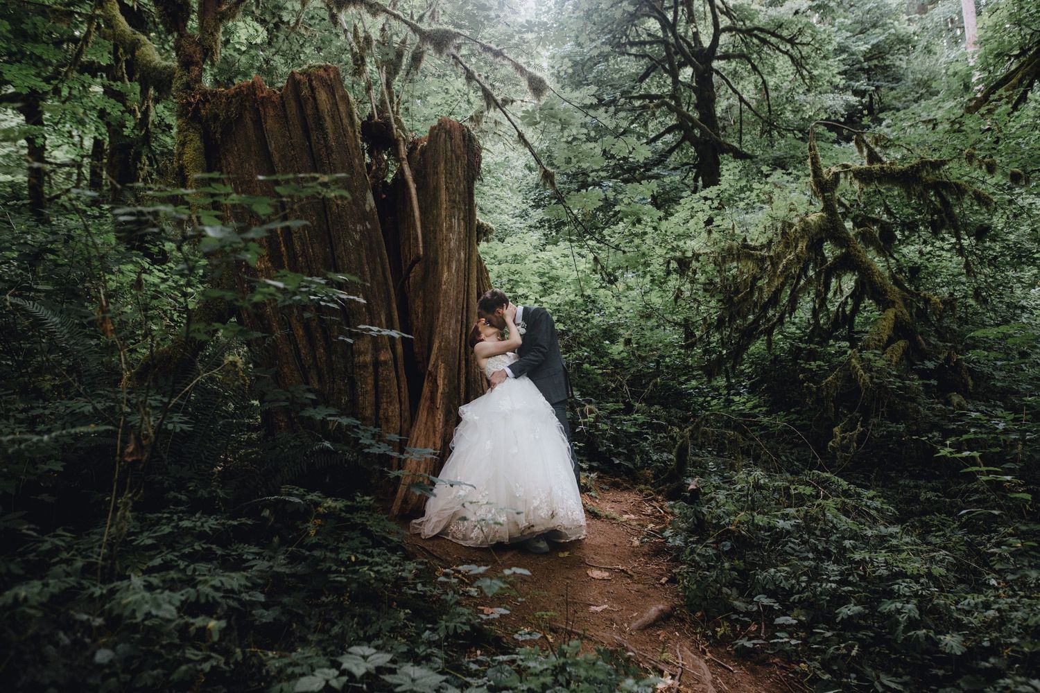 A couple in wedding attire stands together on a wooden bridge surrounded by lush greenery and trees.