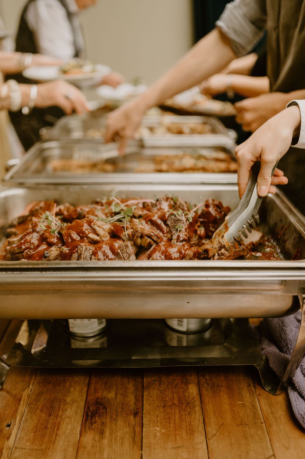 Buffet serving tray filled with saucy meat dish at catered event on wooden floor.