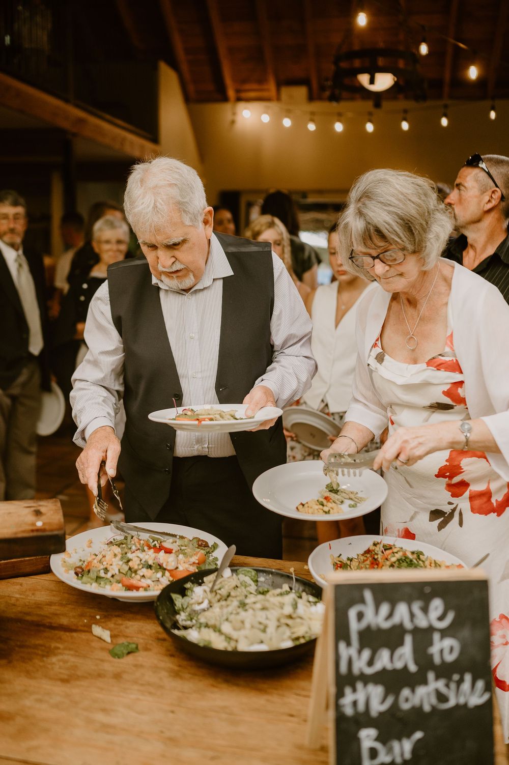 Guests serving themselves from a buffet style meal setup with string lights overhead at an indoor reception.