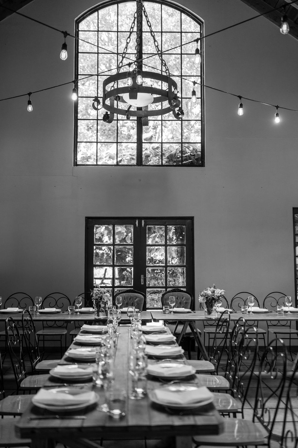 Elegant table setting in a dining room with arched windows, string lights and a rustic chandelier.
