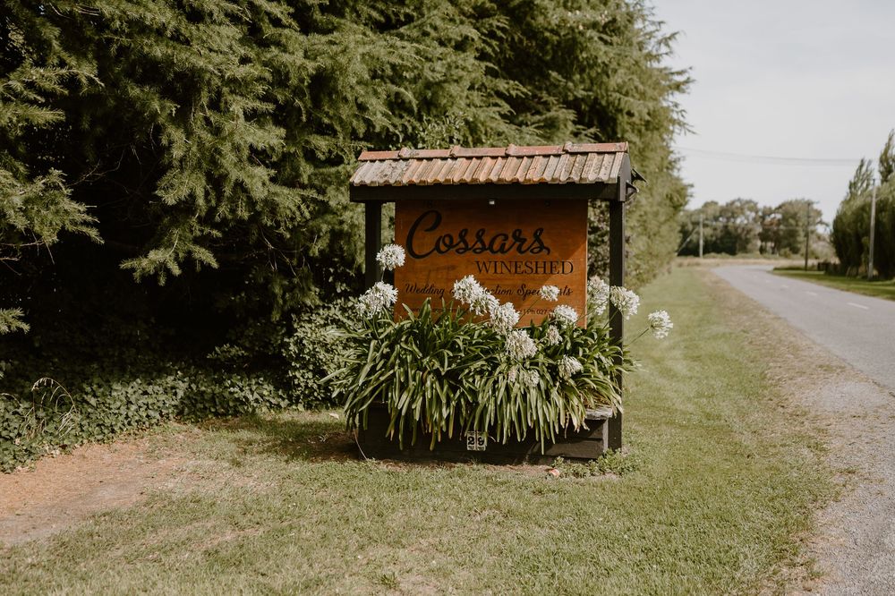 A rustic wooden newspaper box surrounded by lush greenery and flowers along a country road.