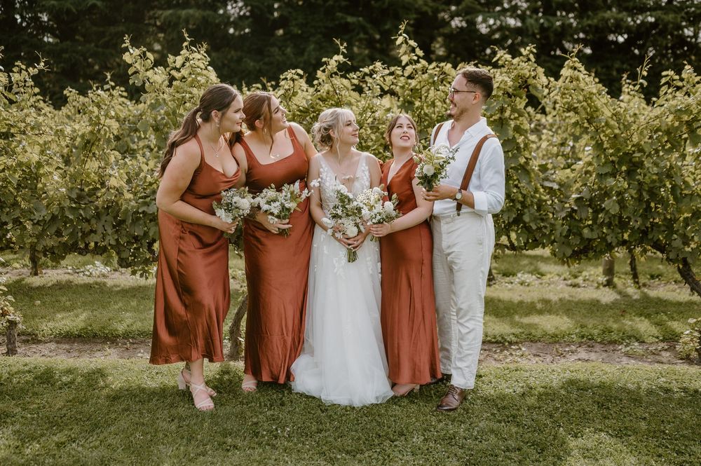 Wedding party in rust-colored dresses poses together on a sunny lawn amid greenery at an outdoor celebration.