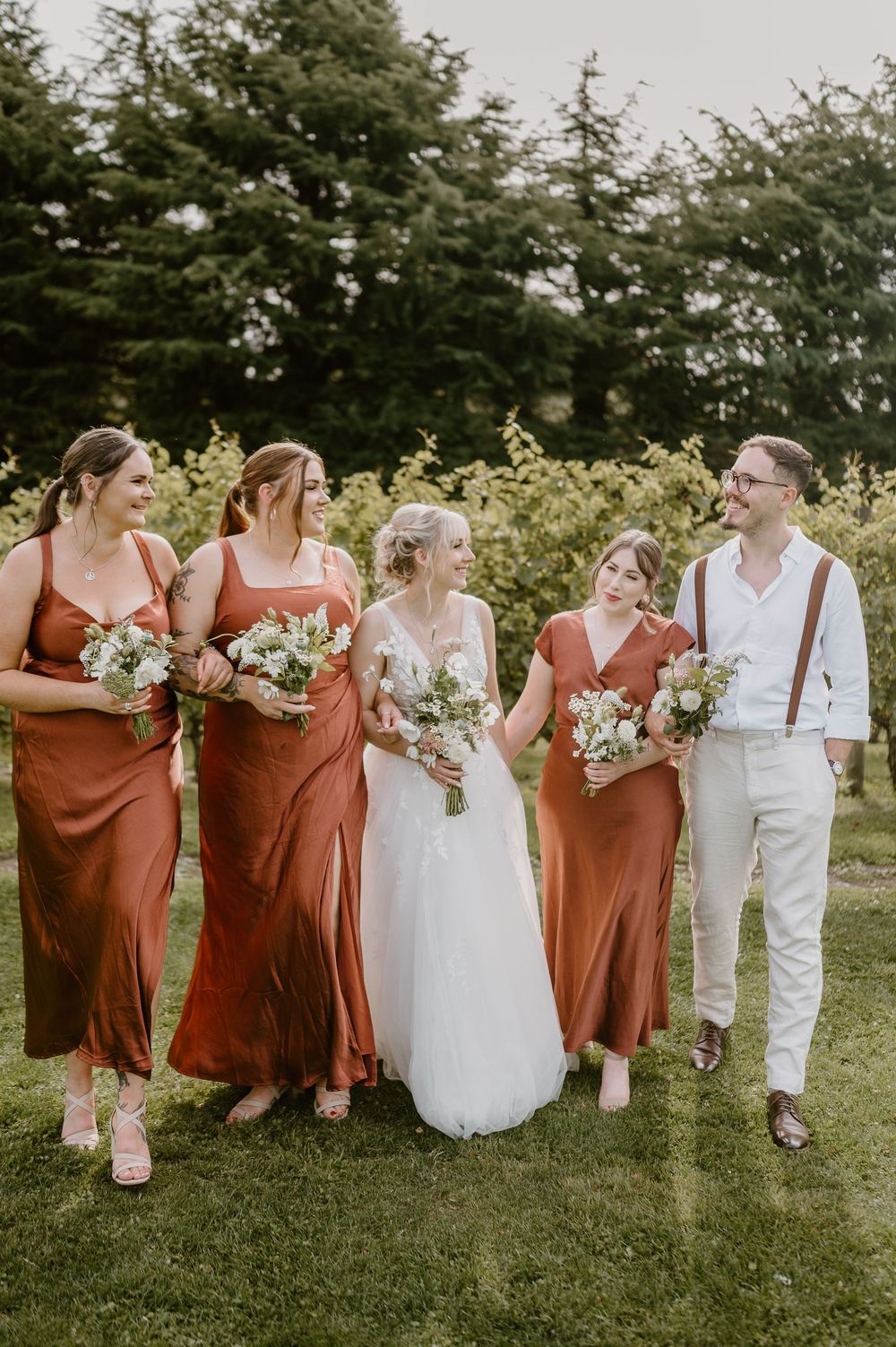 Wedding party in rust-colored bridesmaid dresses and white attire pose together on green lawn with forest backdrop.