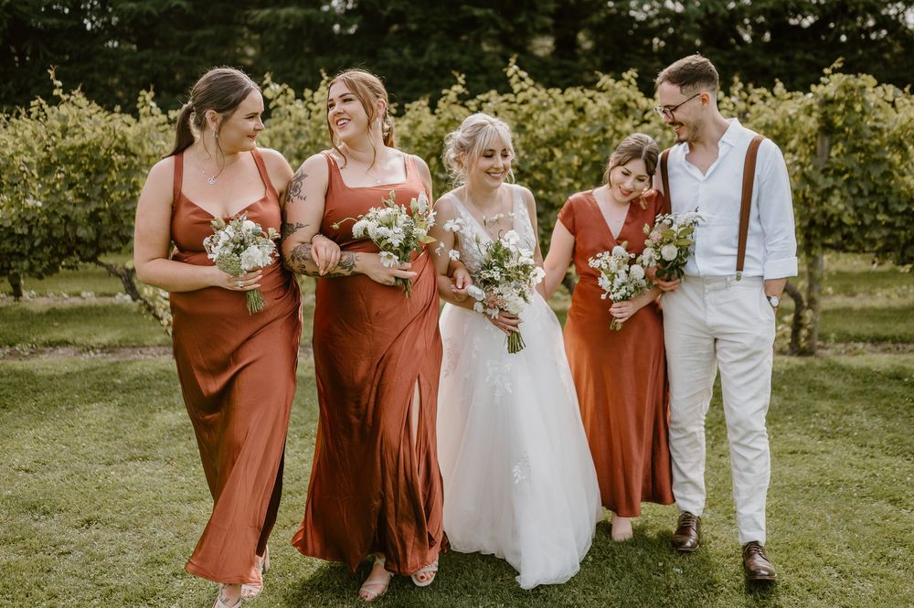 A wedding party in terracotta dresses and white attire walking together outdoors in a garden setting.