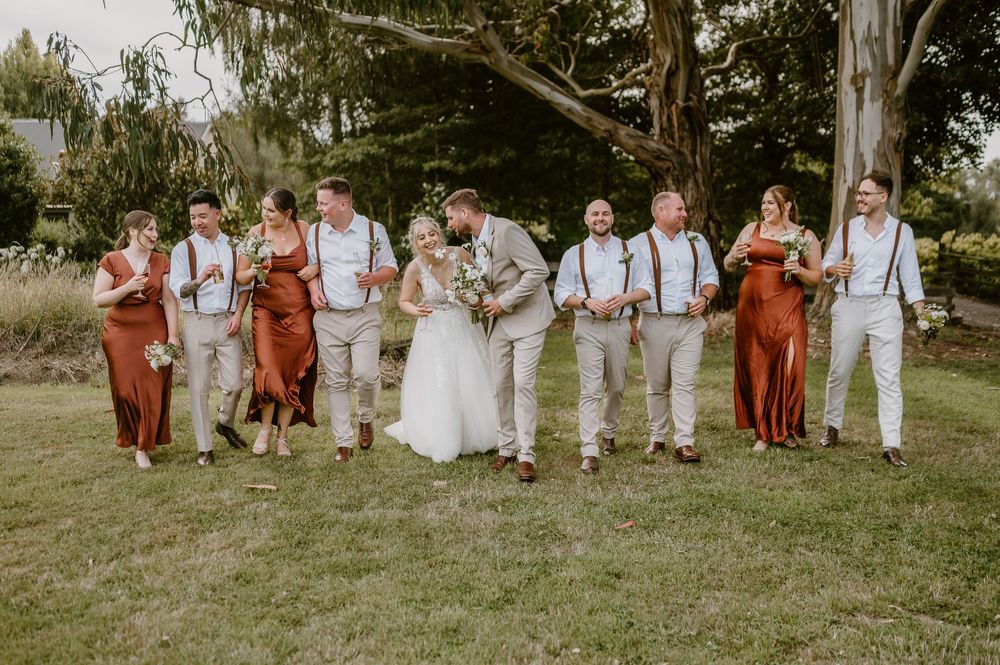 Wedding party in rust-colored dresses and beige suits poses outdoors on grassy field at rustic celebration.