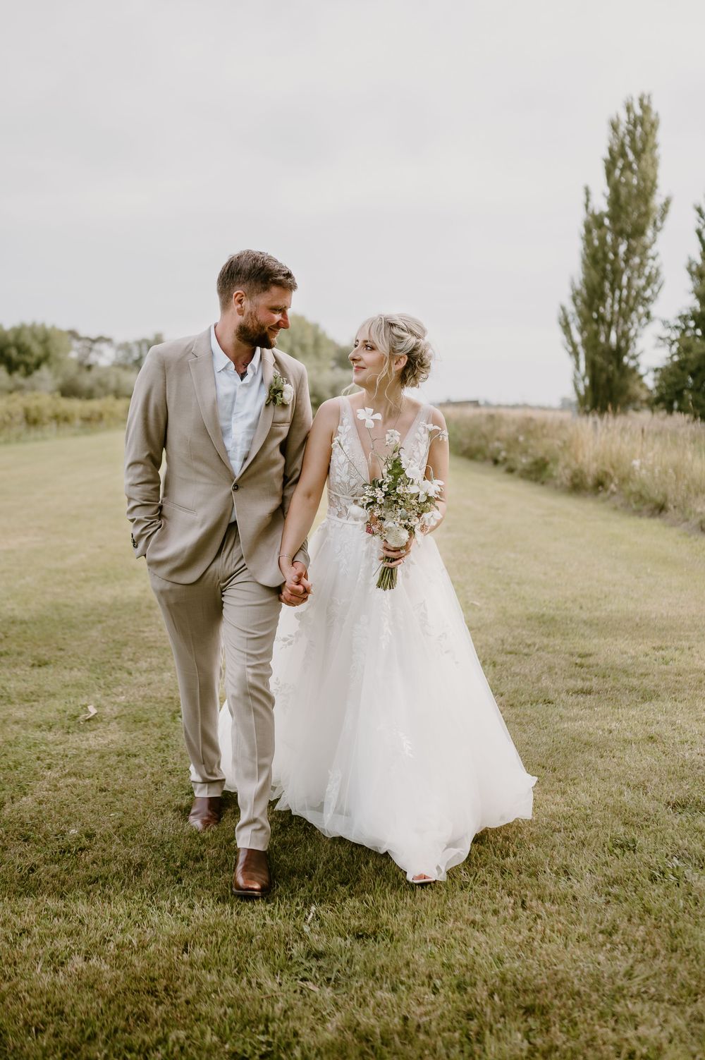 Newlywed couple walking together through a grassy field, bride in white gown and groom in beige suit.