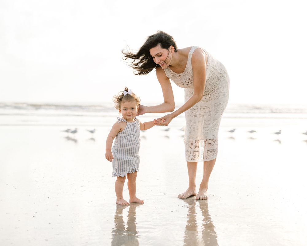 Parent and child hold hands while playing in shallow ocean water.