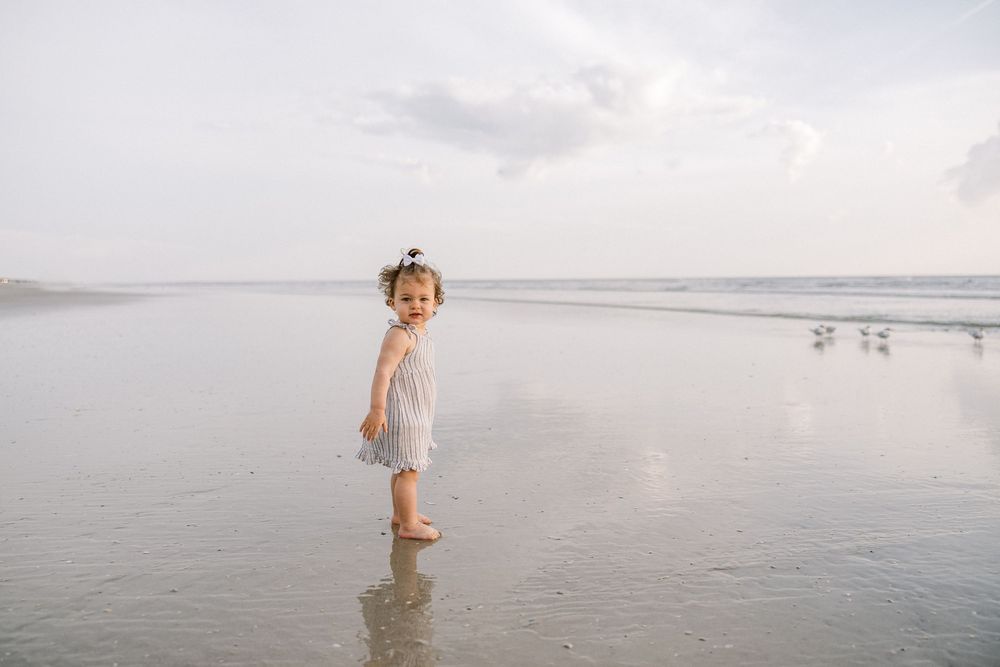 Small child in striped dress stands alone in shallow beach water.