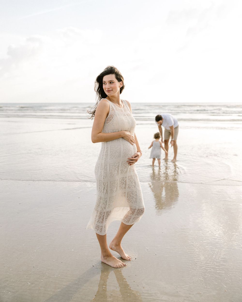 An expectant mother in a white lace dress standing on a serene beach at sunset.