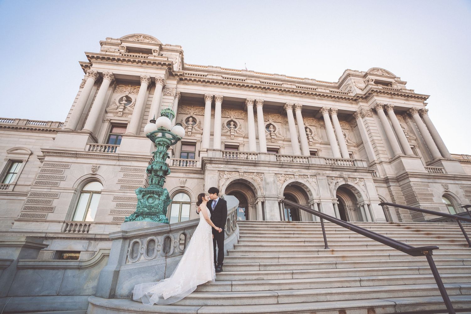 A couple in wedding attire stands on the grand steps of the Library of Congress in Washington DC at dusk.