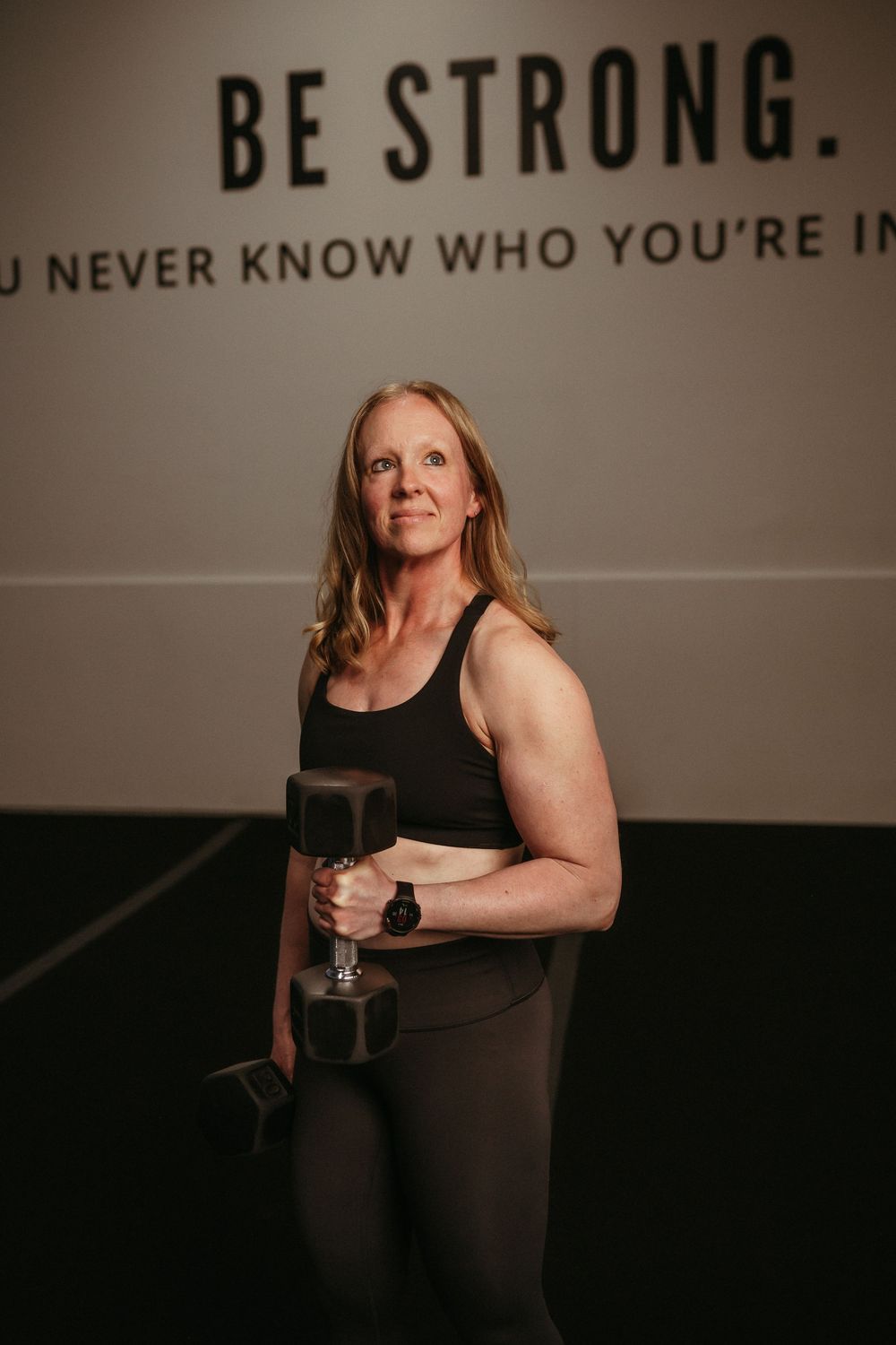 A woman in workout attire holds a dumbbell while standing in a gym with 'BE STRONG' text on the wall.