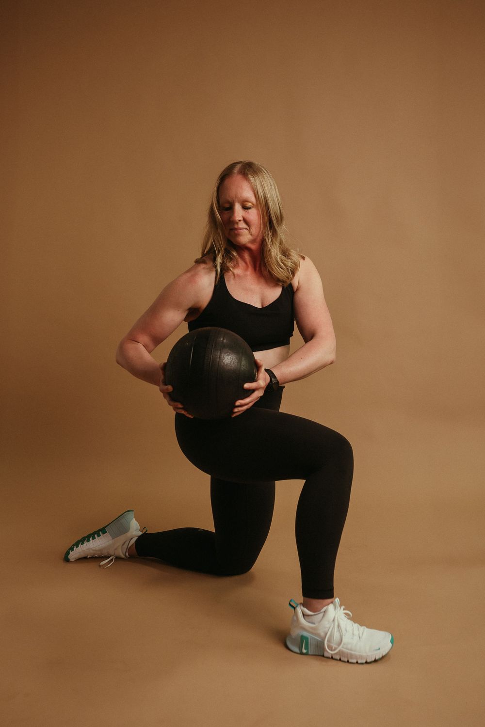 Athletic person in black workout attire performs a lunge exercise with a medicine ball against brown background.