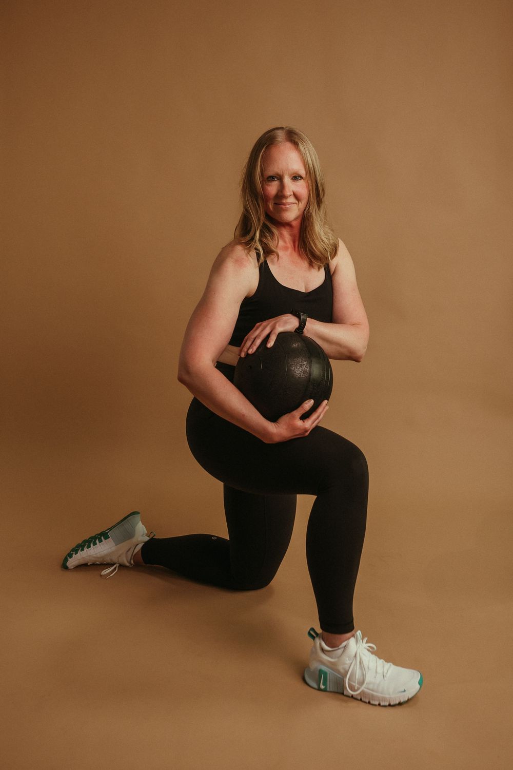 Person in black workout attire kneels while holding a heavy medicine ball against brown background.
