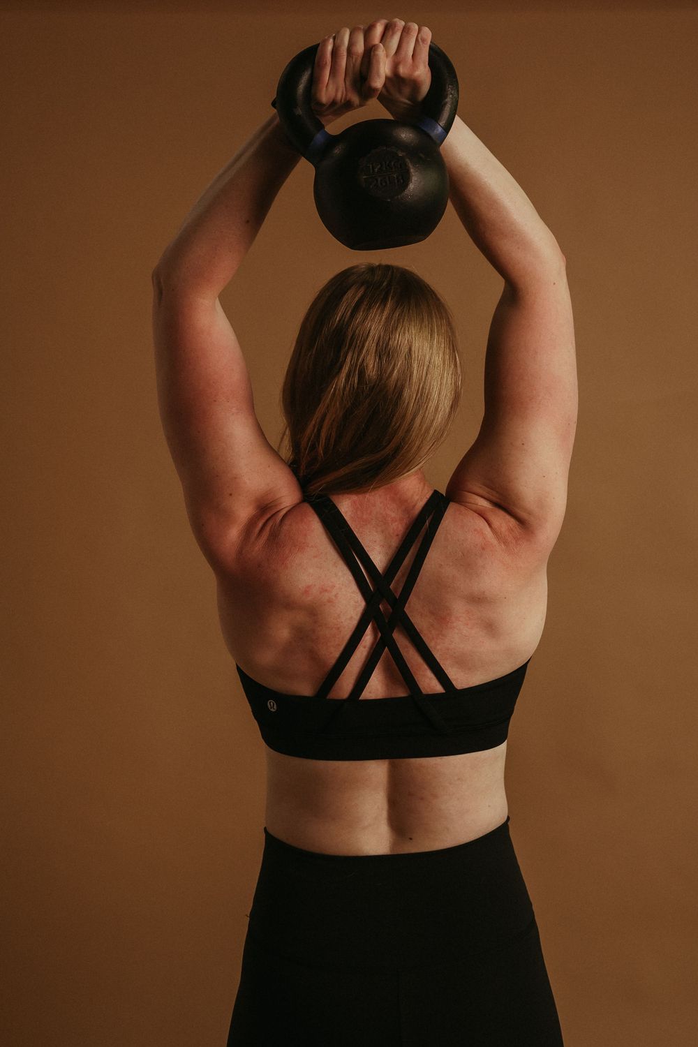 Athletic person in black workout wear holds a kettlebell overhead against brown background during strength training exercise.