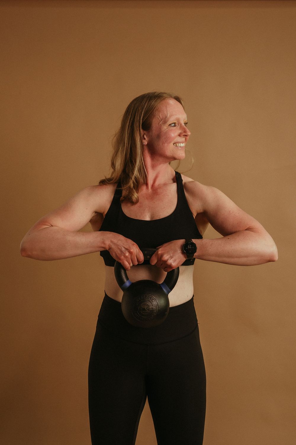 Person in black athletic wear demonstrates proper form for a kettlebell workout against warm brown backdrop.